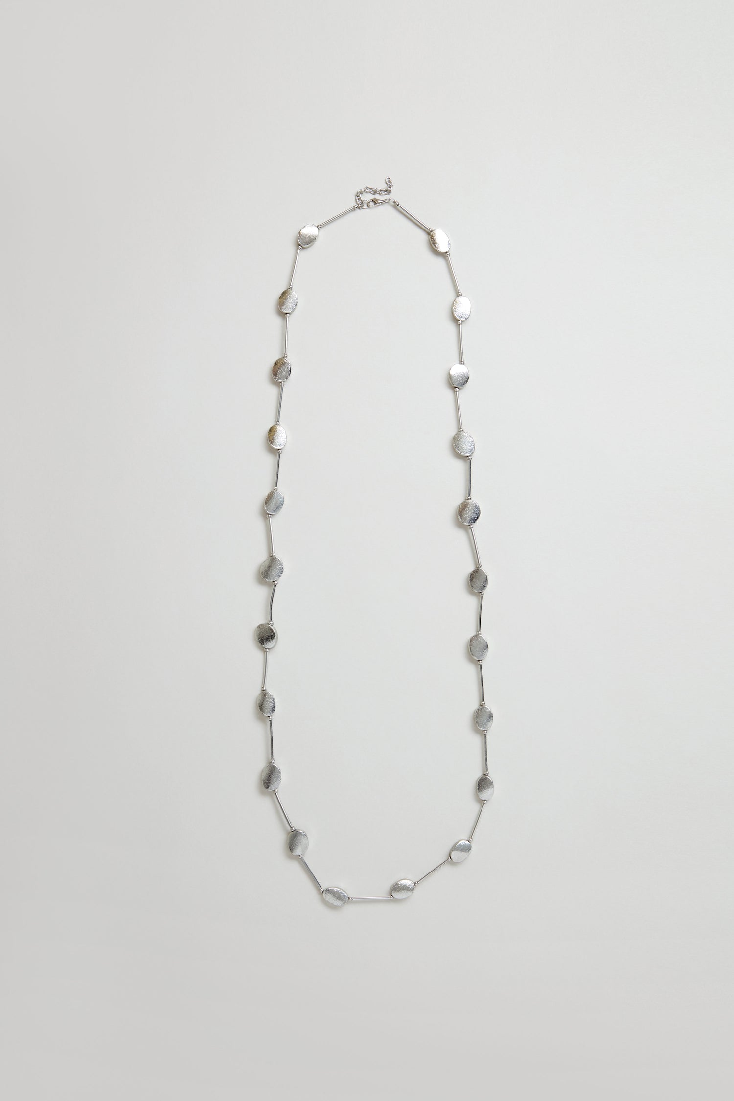 The Long Oval Disc Necklace features textured silver oval discs evenly spaced along a thin chain, shown against a plain light background.