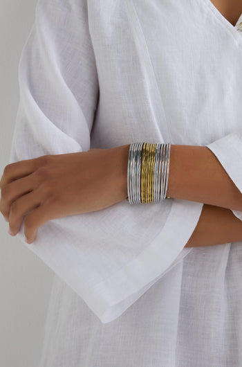 A person in a white linen top crosses their arm, highlighting the Mixed Metal Cuff—a wide, hammered bracelet in silver and gold tones that makes a bold statement.