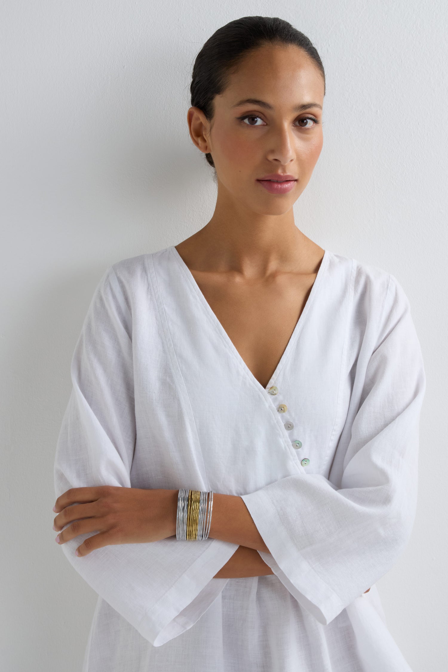 A woman with medium skin tone stands against a plain white wall, arms lightly crossed, wearing a white long-sleeved V-neck top and the Mixed Metal Cuff—a hammered-finish bracelet.