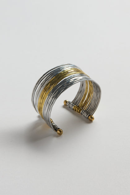 The Mixed Metal Cuff features hammered silver and gold-toned textured bands, open ends with round finials, and is styled as a versatile statement piece on a plain white surface.