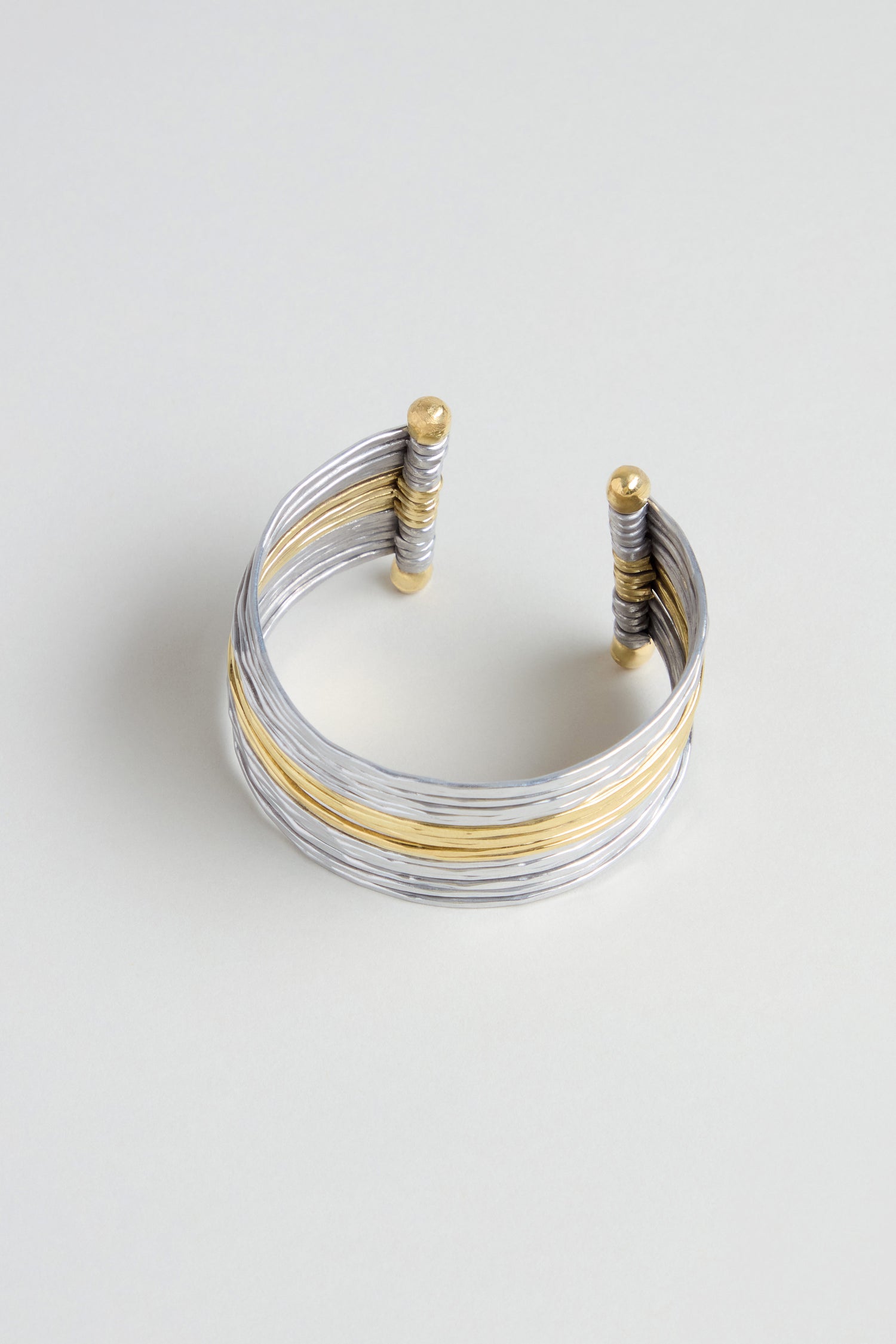 The Mixed Metal Cuff showcases silver and gold wrapped strands with gold-accented ends, creating a striking mixed metal bracelet displayed on a light background.