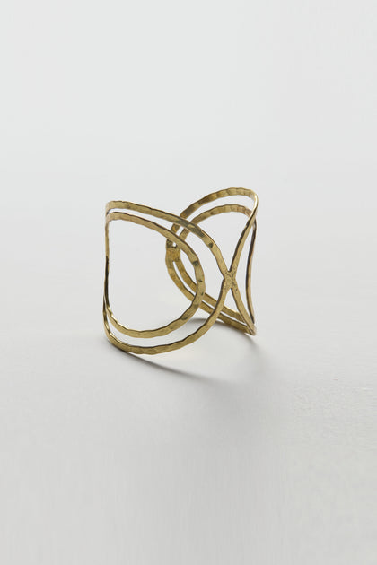 The Butterfly Bangle is a gold cuff-style bracelet with a hammered texture, featuring an intersecting crisscross design and sculptural silhouette, displayed on a plain white background.