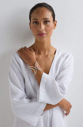 A woman in a white, loose-fitting garment stands against a white wall, wearing the modern Butterfly Bangle on her left wrist and looking directly at the camera.