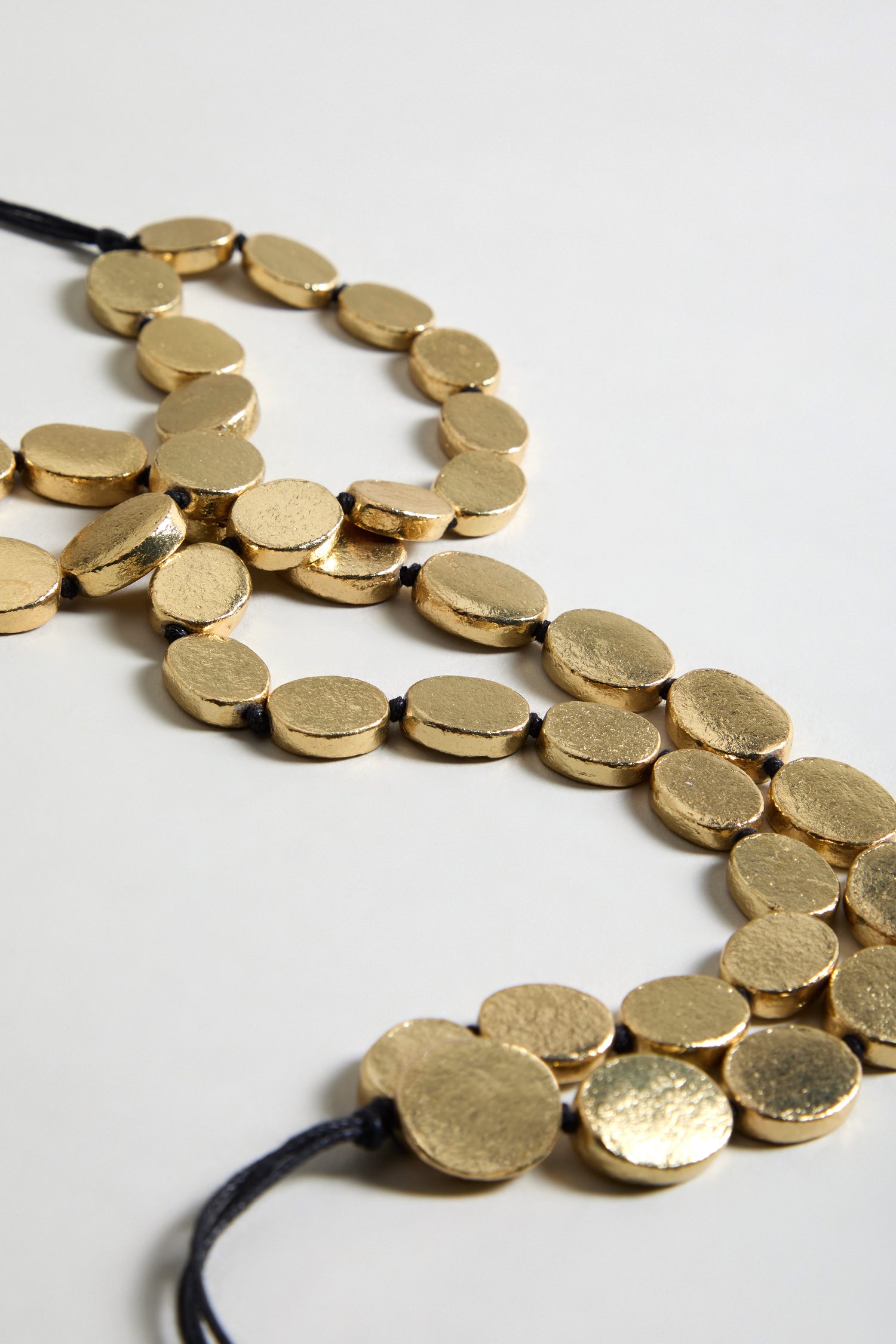 The Double Strand Oval Disc Necklace features gold-colored, textured oval discs on a black cord, arranged in a loose curve on a neutral background.