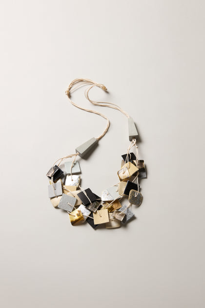The Gathered Shapes Necklace showcases irregular metallic squares and rectangles in silver, gold, and black, strung on a light-toned cord for a modern look.