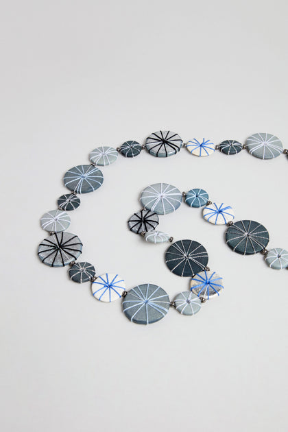 The Long Wooden Metallic Parasols Necklace features round, flat beads in various sizes with geometric line patterns in blue, gray, black, and white, arranged spirally on a plain background.