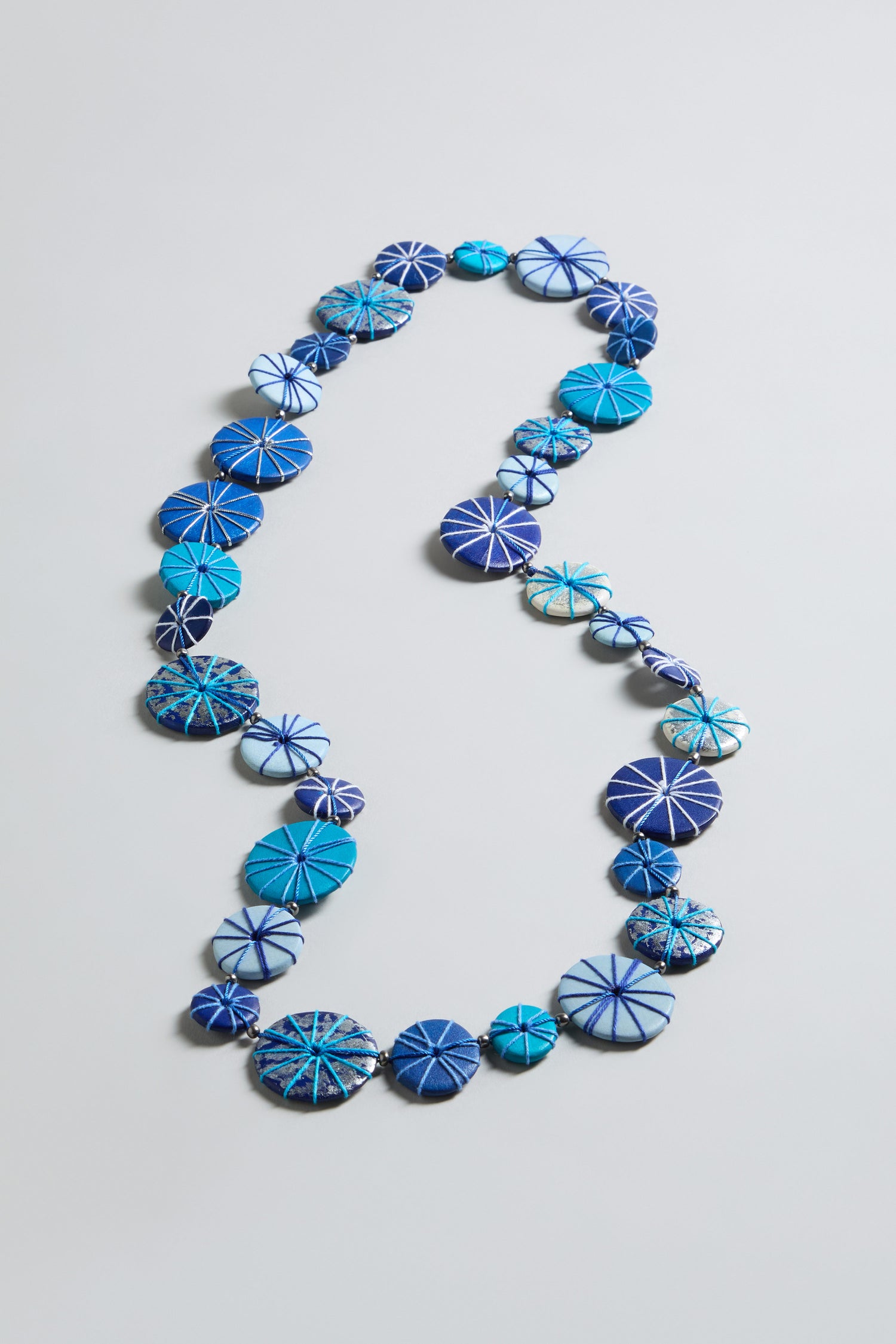 The Long Wooden Disc Necklace showcases flat, round blue beads with white radial patterns, beautifully arranged on a simple light background.