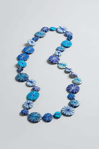 The Long Wooden Disc Necklace showcases flat, round blue beads with white radial patterns, beautifully arranged on a simple light background.
