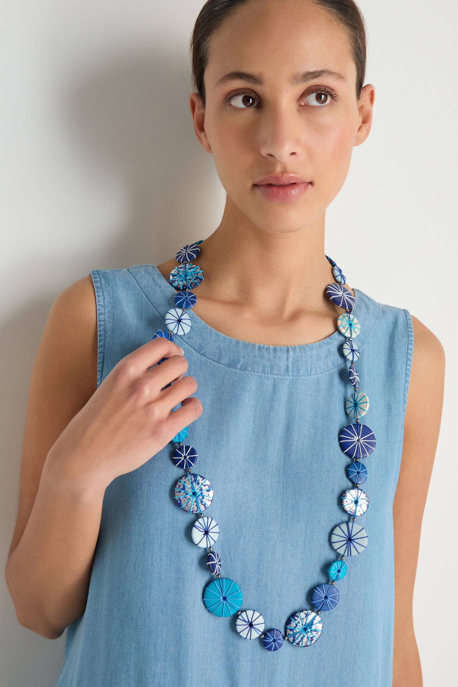 A woman in a sleeveless light blue denim dress wears the Long Wooden Disc Necklace featuring patterned blue and white wooden discs with thread wrap detail, looking slightly to the side.