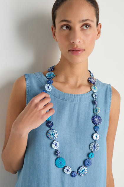 A woman in a sleeveless light blue denim dress wears the Long Wooden Disc Necklace featuring patterned blue and white wooden discs with thread wrap detail, looking slightly to the side.