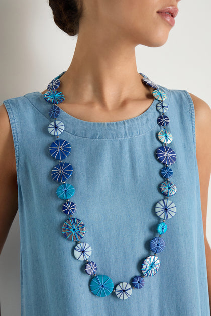 A woman in a sleeveless light blue dress showcases the Long Wooden Disc Necklace, which features blue and white patterned beads, an adjustable cord, and thread wrap detail.