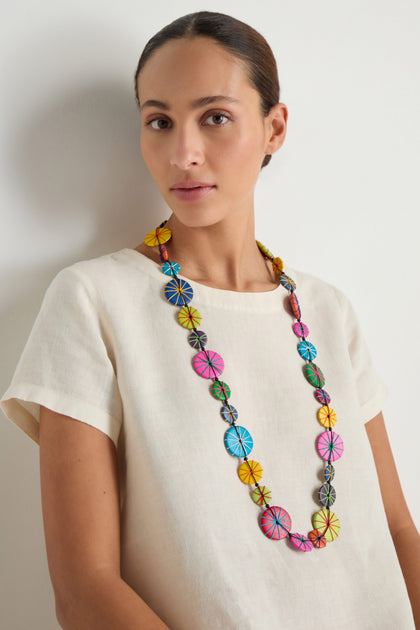 A woman stands against a plain light background wearing a cream short-sleeve top and the Long Wooden Disc Necklace—an adjustable cord necklace with circular, multicolored wooden beads.