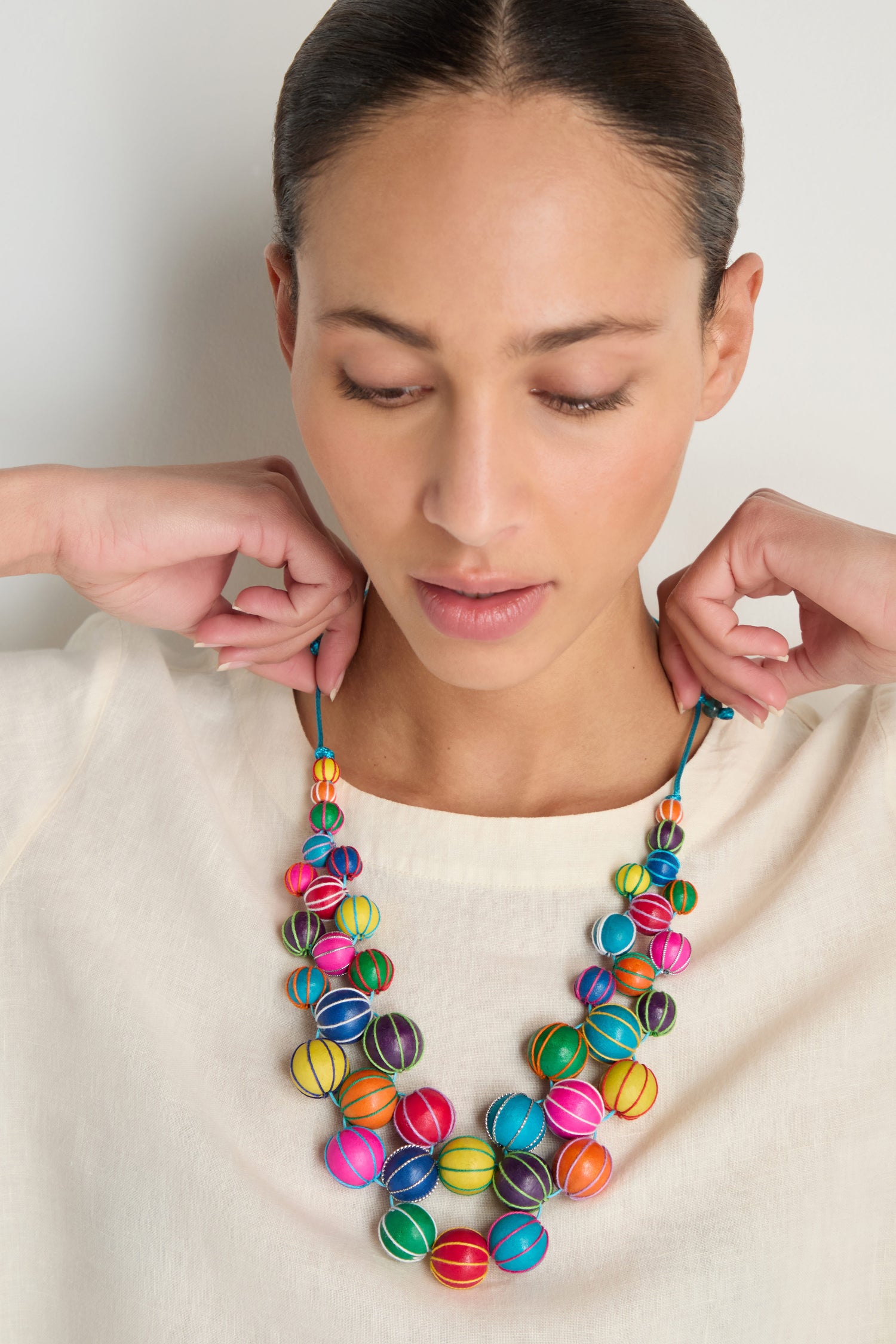 A person in a cream top adjusts the Rainbow Wooden Spheres Necklace, which features round beads in vibrant rainbow colors.