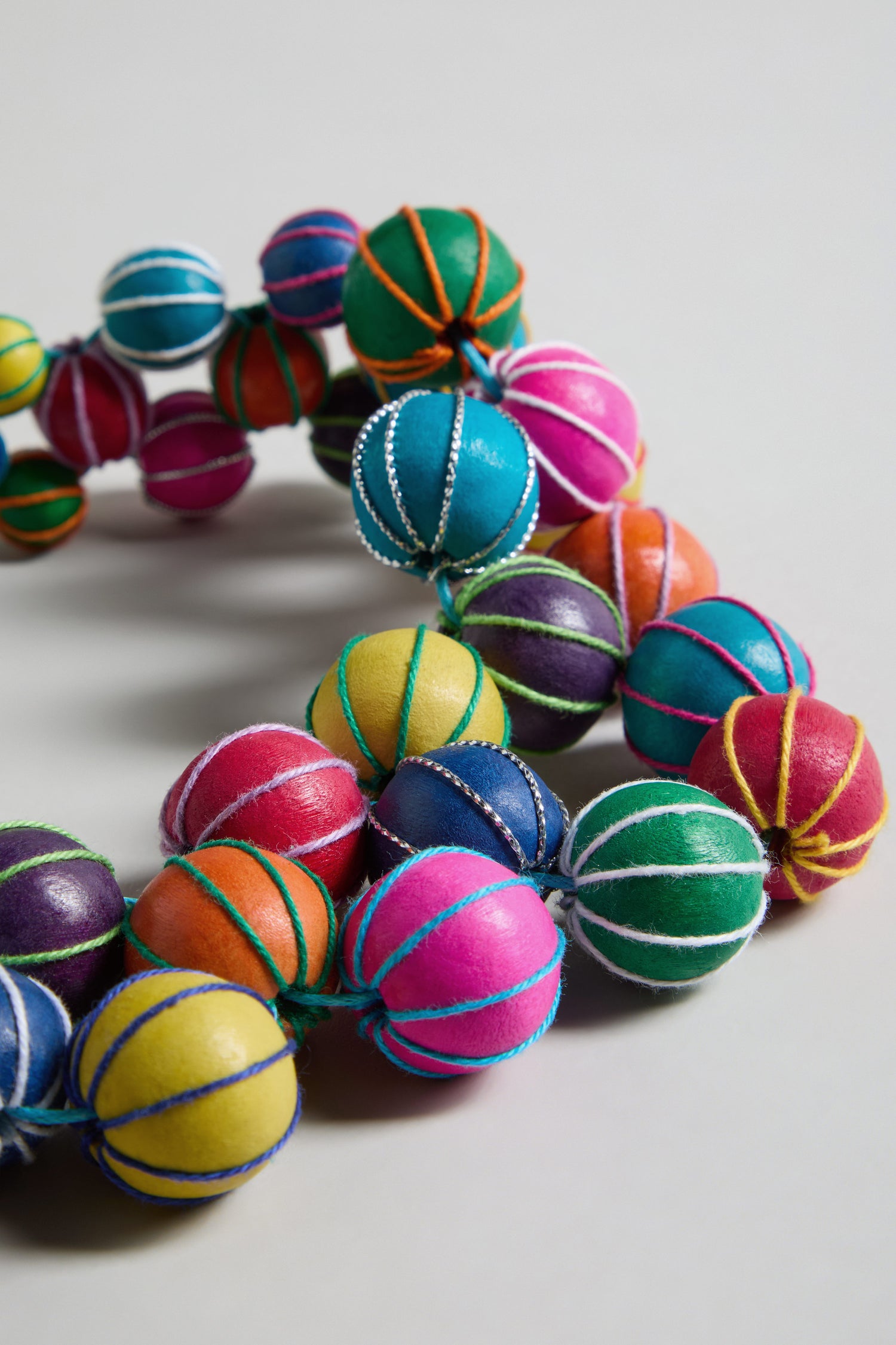 The Rainbow Wooden Spheres Necklace features vibrant wooden beads, each wrapped with contrasting thread, displayed in a close-up on a light background.