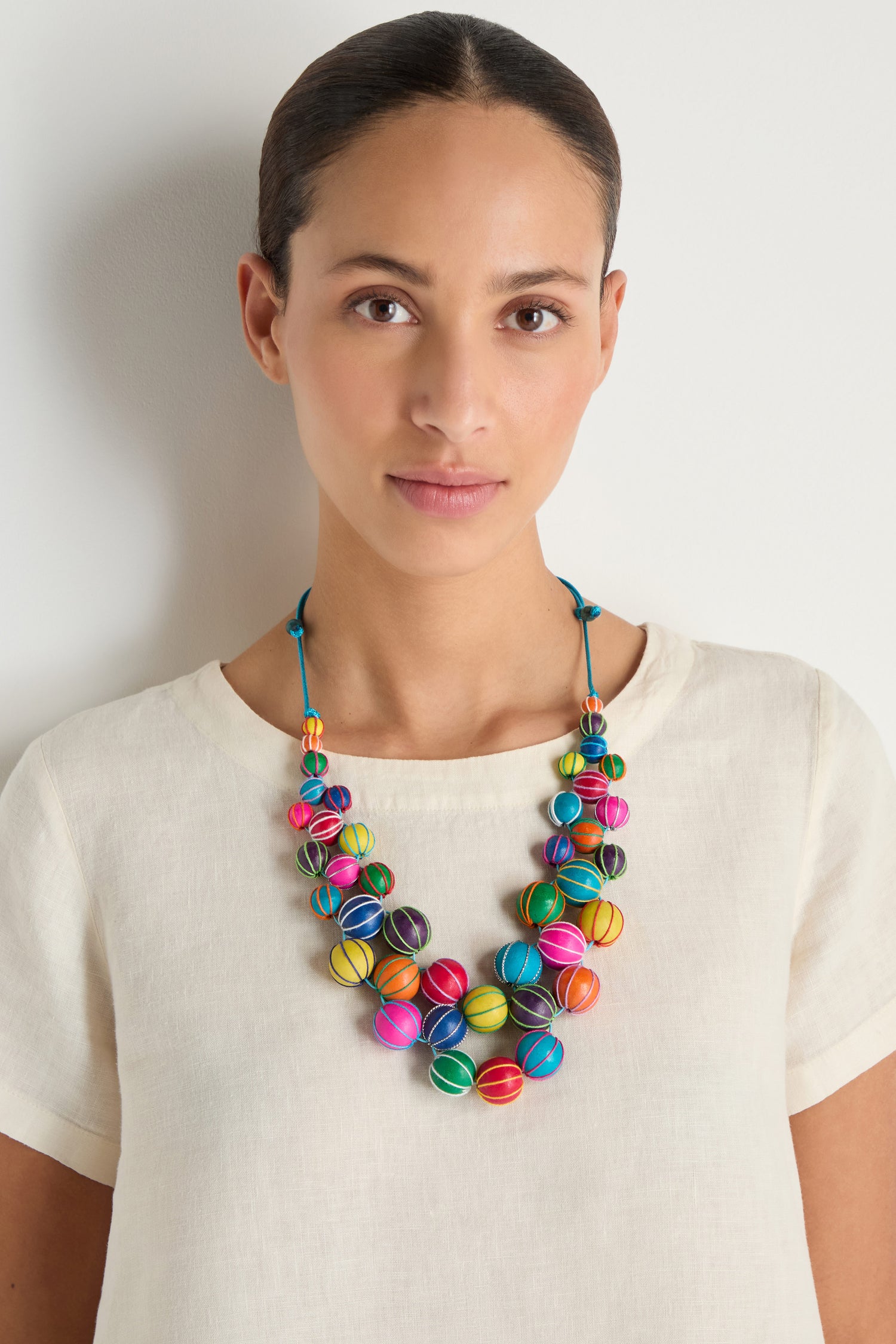 A woman stands against a plain white background, looking at the camera while wearing a cream-colored top and the Rainbow Wooden Spheres Necklace.