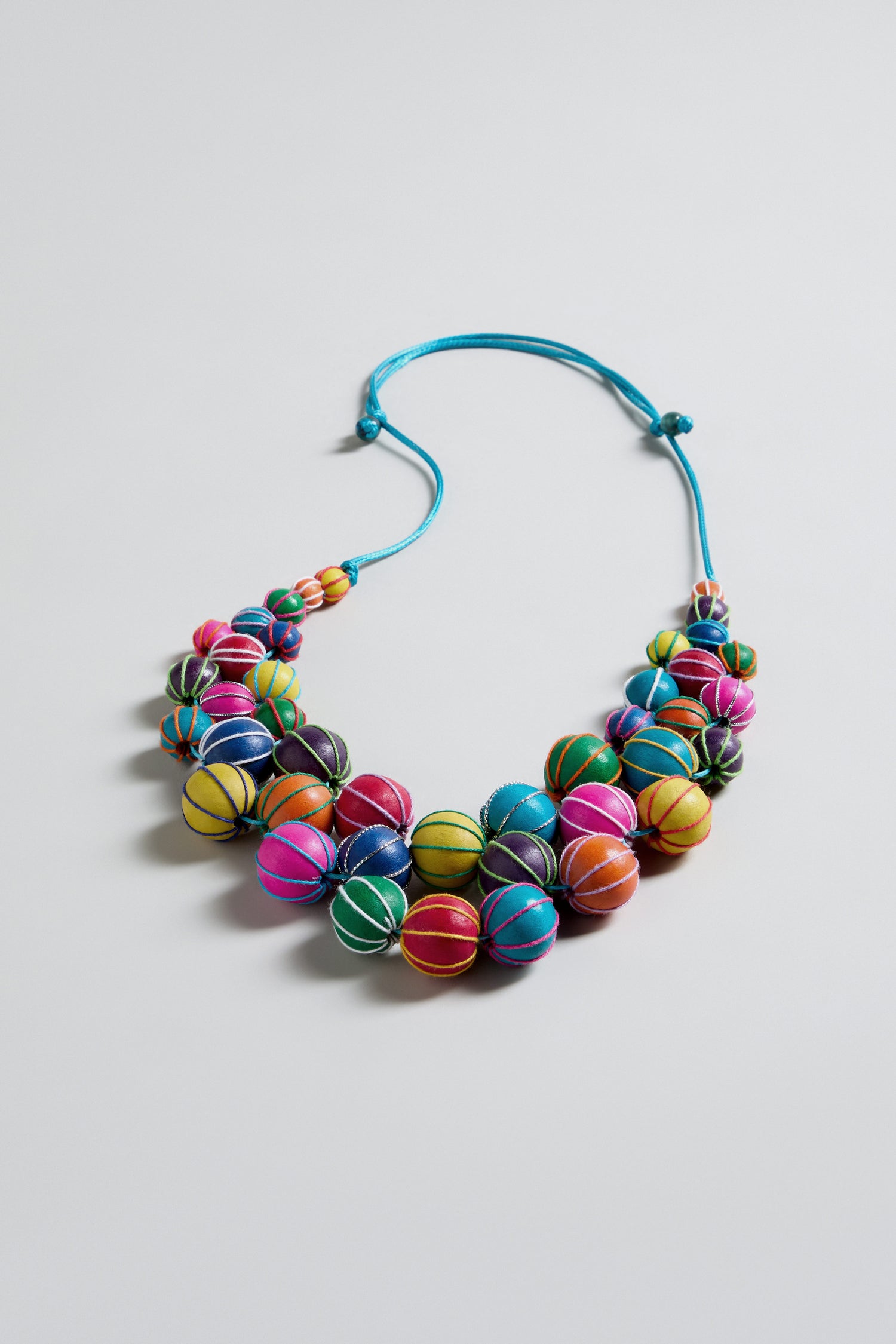 The Rainbow Wooden Spheres Necklace features rainbow-hued wooden beads, each wrapped with thread and strung on a light blue adjustable cord, shown against a plain white background.