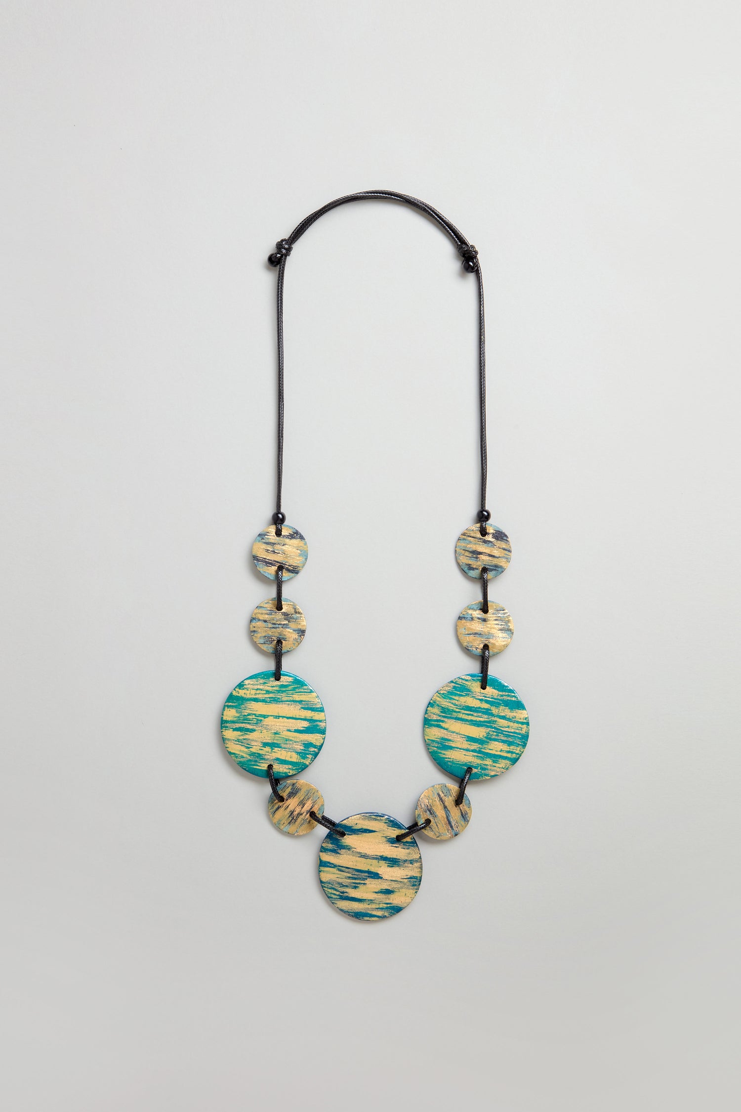 The Painted Disc Necklace features seven round, flat beads in blue and beige patterns on a black adjustable cord, shown against a plain light background.