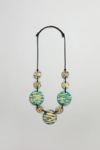 The Painted Disc Necklace features seven round, flat beads in blue and beige patterns on a black adjustable cord, shown against a plain light background.