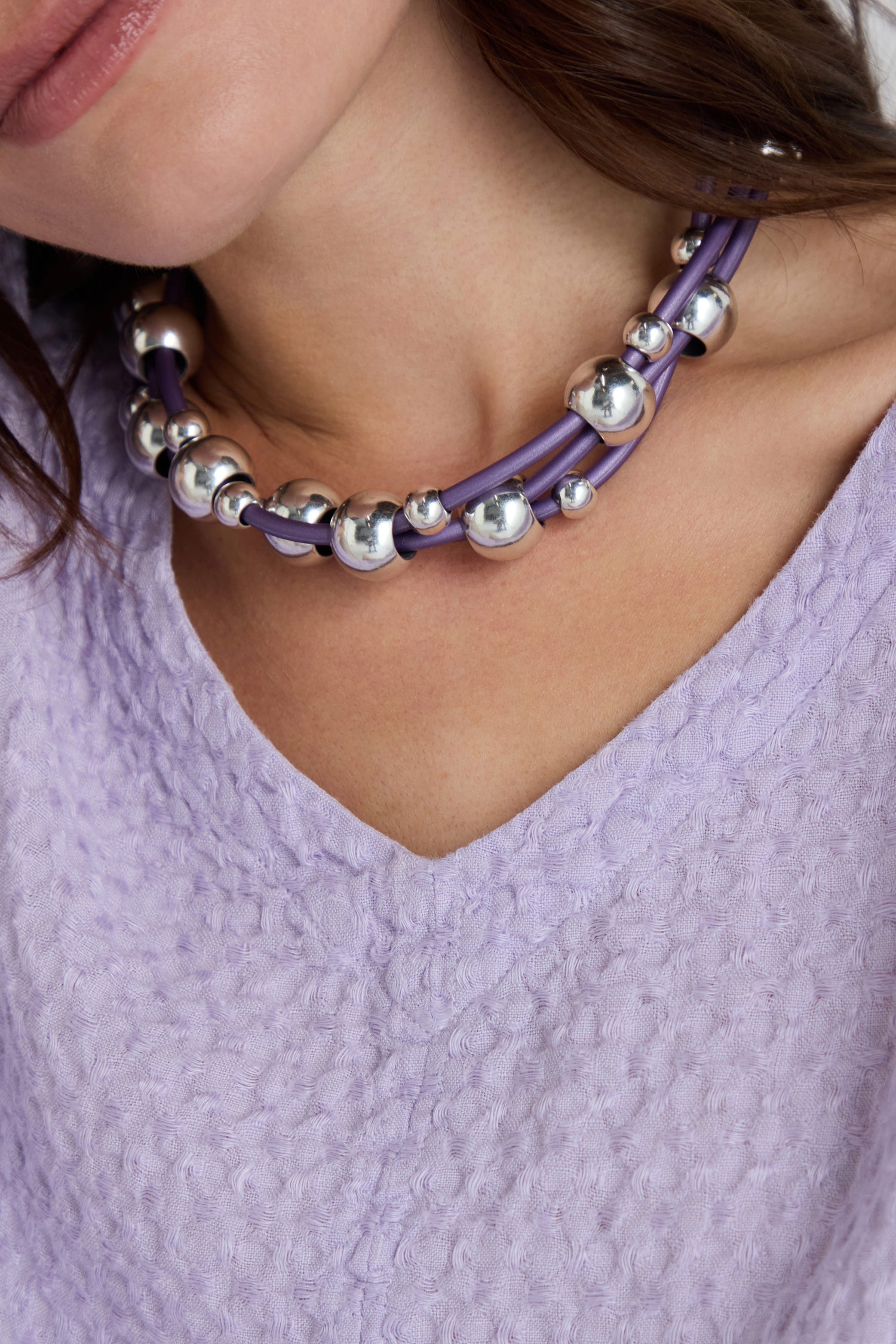 A woman wears a textured lavender top and the Infinite Spheres Necklace—a chunky silver acrylic piece featuring purple cords and a secure magnetic clasp.