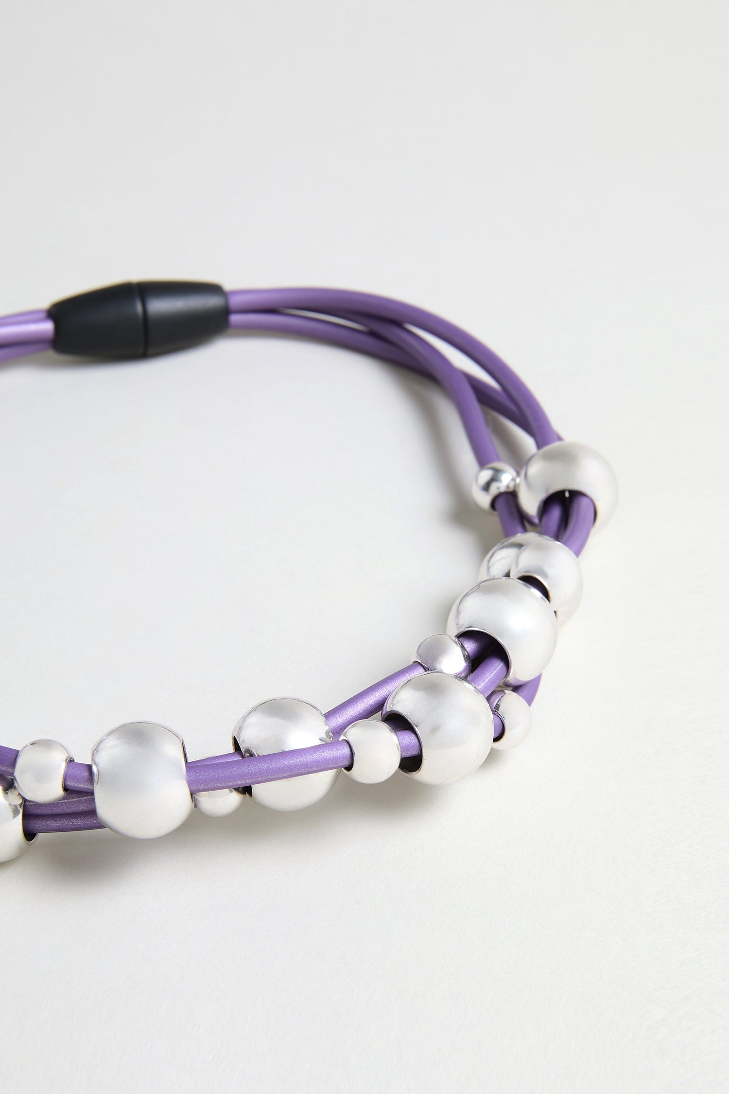 The Infinite Spheres Necklace showcases violet PVC cords entwined with silver acrylic spheres in assorted sizes, set against a simple light background.