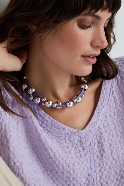 Woman in a textured lavender top wears the Infinite Spheres Necklace, which features silver acrylic spheres on violet PVC cords, as she looks down with a relaxed expression.