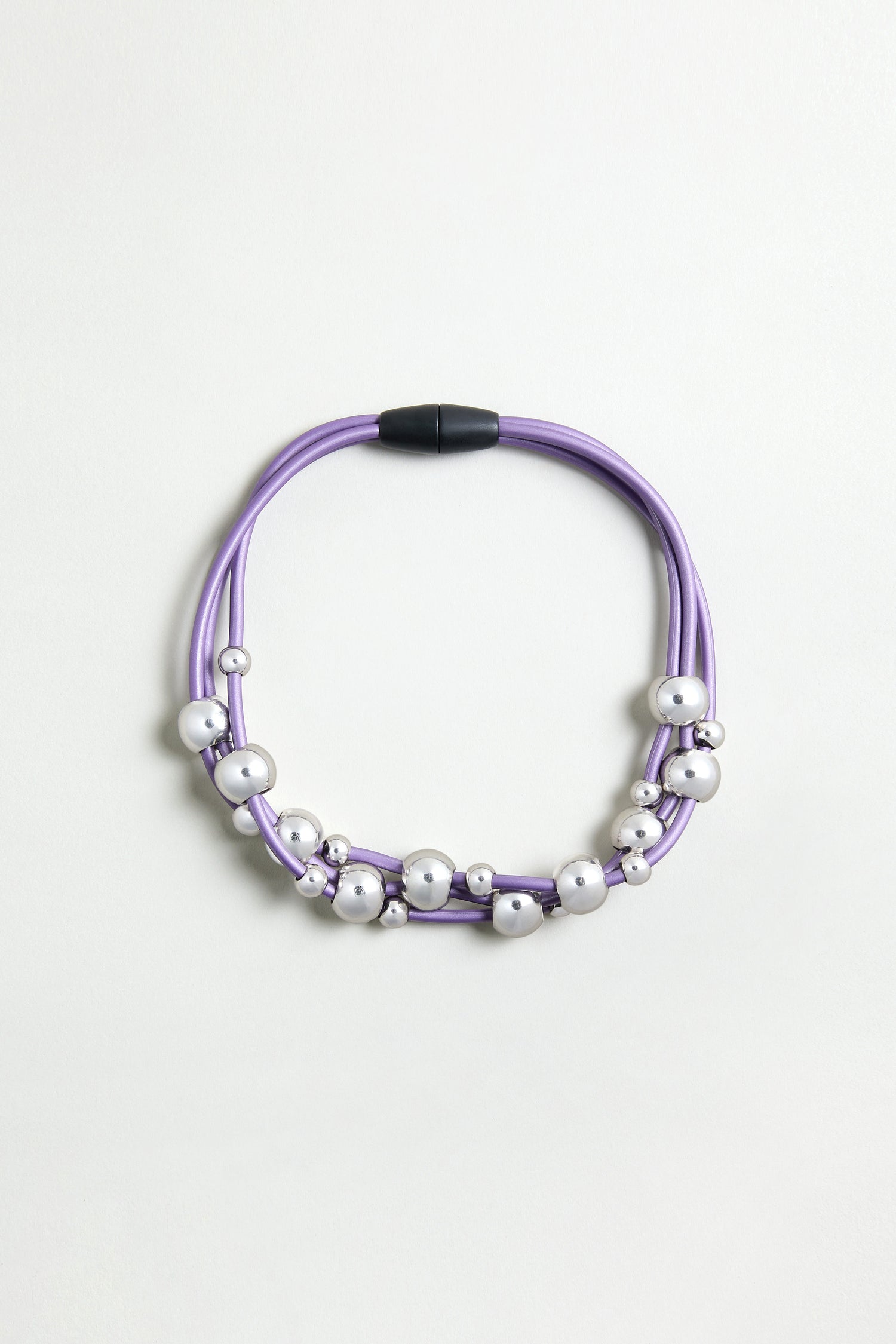 The Infinite Spheres Necklace has violet PVC cords, silver acrylic spheres, and a black magnetic clasp, arranged circularly on a light background.