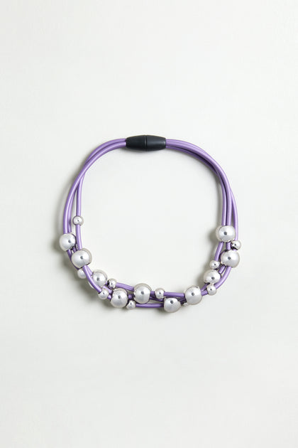 The Infinite Spheres Necklace has violet PVC cords, silver acrylic spheres, and a black magnetic clasp, arranged circularly on a light background.