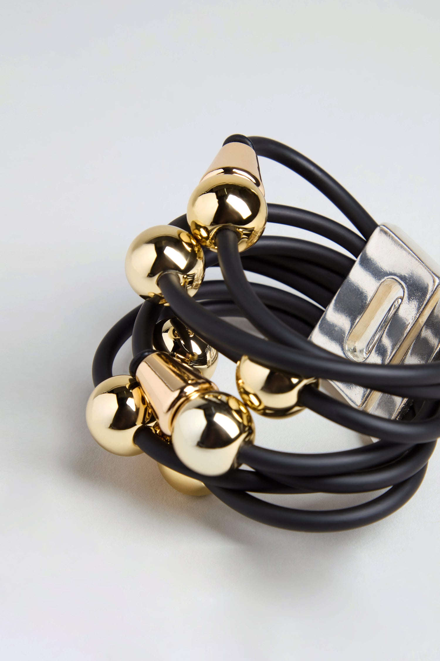 The Metal Spheres Bracelet features multiple black elastic bands adorned with sculptural shiny gold spheres and finished with a metallic clasp, displayed on a light surface.