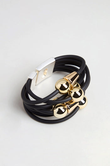 The Metal Spheres Bracelet is a black multi-strand bracelet accented with gold-tone metal beads and finished with a rectangular silver-tone magnetic clasp. Featuring a sculptural design, this handmade piece from France is showcased on a neutral background.