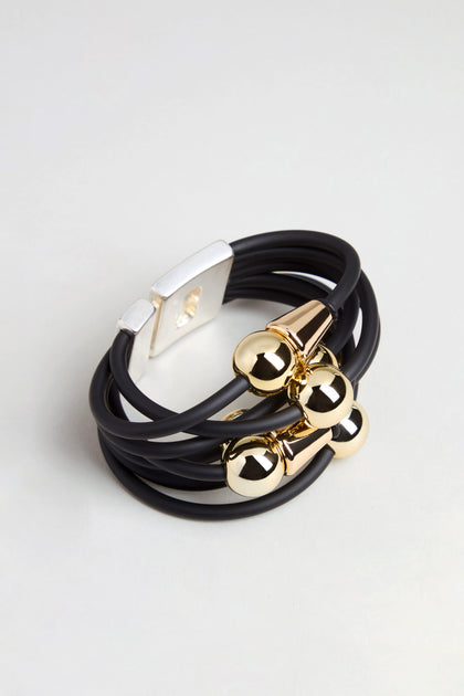 The Metal Spheres Bracelet is a black multi-strand bracelet accented with gold-tone metal beads and finished with a rectangular silver-tone magnetic clasp. Featuring a sculptural design, this handmade piece from France is showcased on a neutral background.