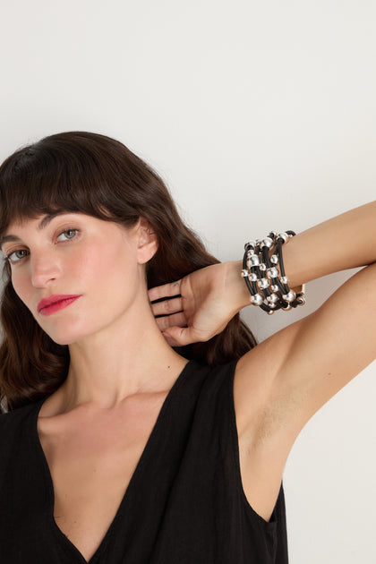 A woman with brown hair wearing a black sleeveless top poses with one arm raised, showcasing an array of black and silver beaded bracelets—including the standout Metal Spheres Bracelet, known for its sculptural design—on her wrist against a plain white background. Each piece is handmade in France, lending an artisanal touch to her look.