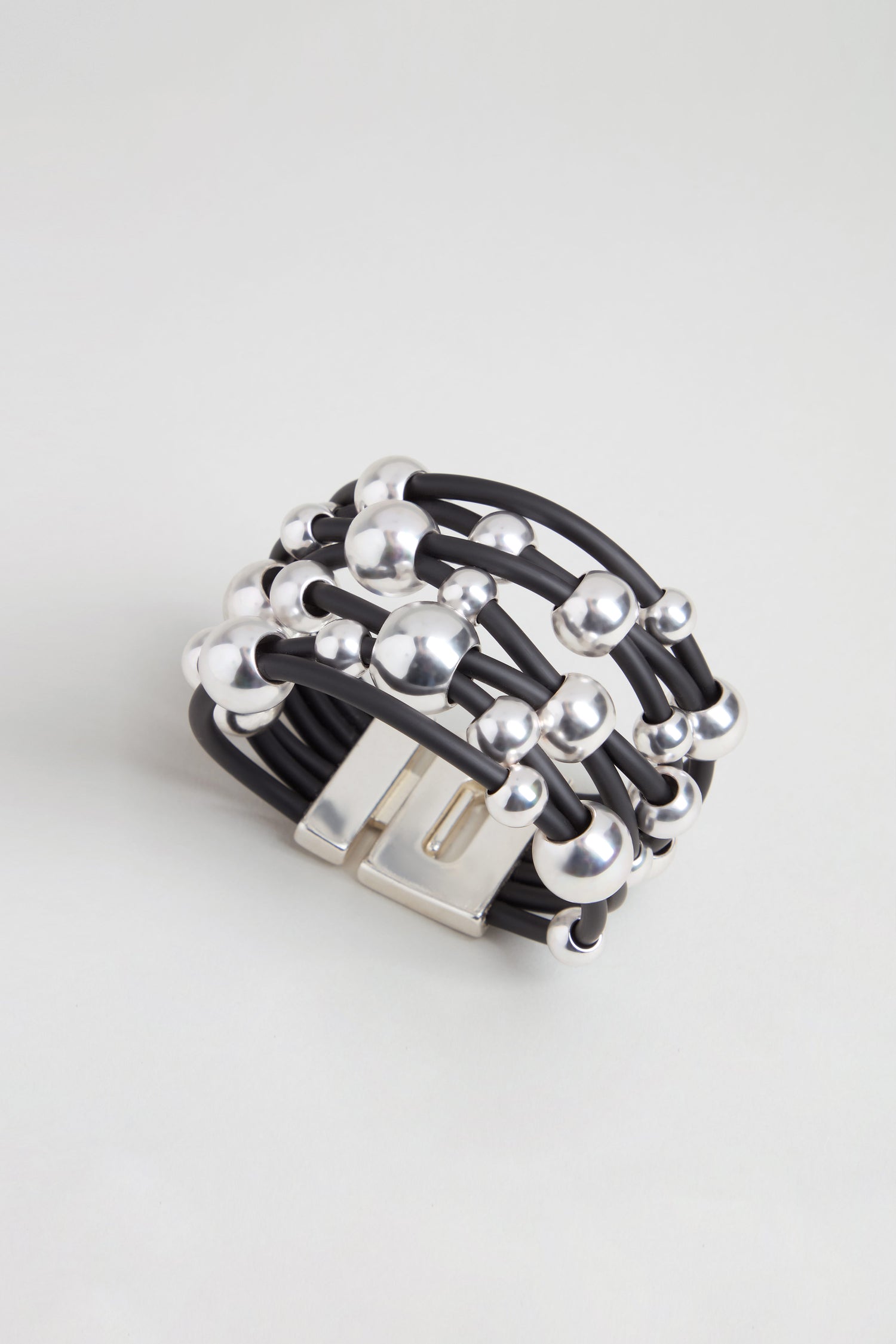 The Metal Spheres Bracelet features a black multi-strand design accented with silver beads and a rectangular silver clasp, showcasing a sculptural aesthetic and elegantly displayed on a light gray background.