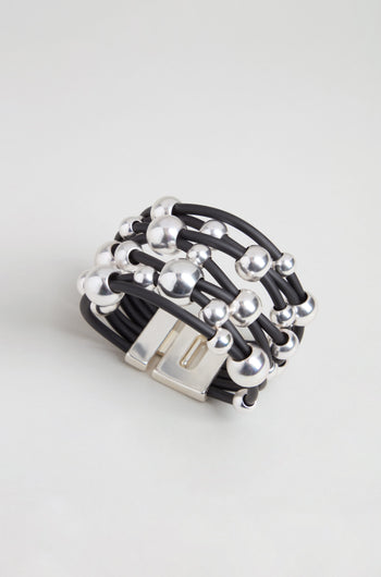 The Metal Spheres Bracelet features a black multi-strand design accented with silver beads and a rectangular silver clasp, showcasing a sculptural aesthetic and elegantly displayed on a light gray background.