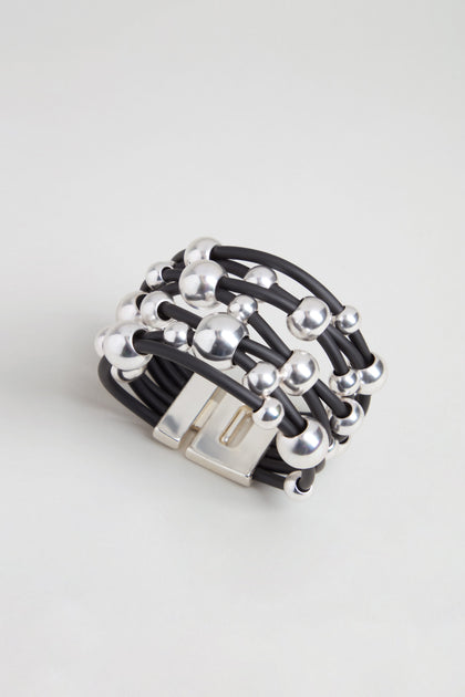 The Metal Spheres Bracelet features a black multi-strand design accented with silver beads and a rectangular silver clasp, showcasing a sculptural aesthetic and elegantly displayed on a light gray background.