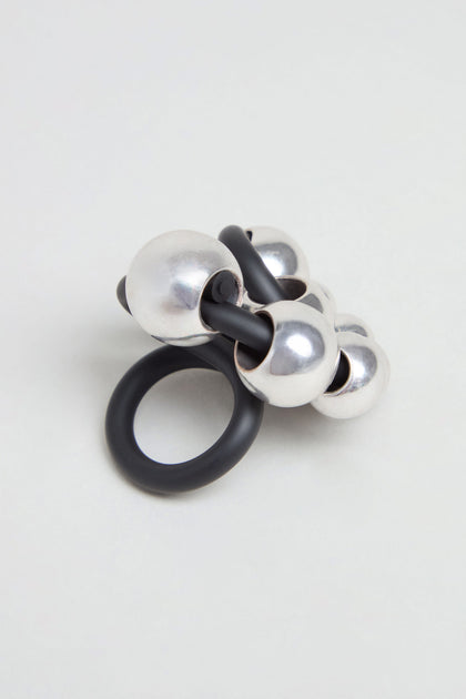 The Metal Spheres Adjustable Ring showcases a sculptural design with interconnected black and silver metallic spheres, elegantly displayed on a plain light background.
