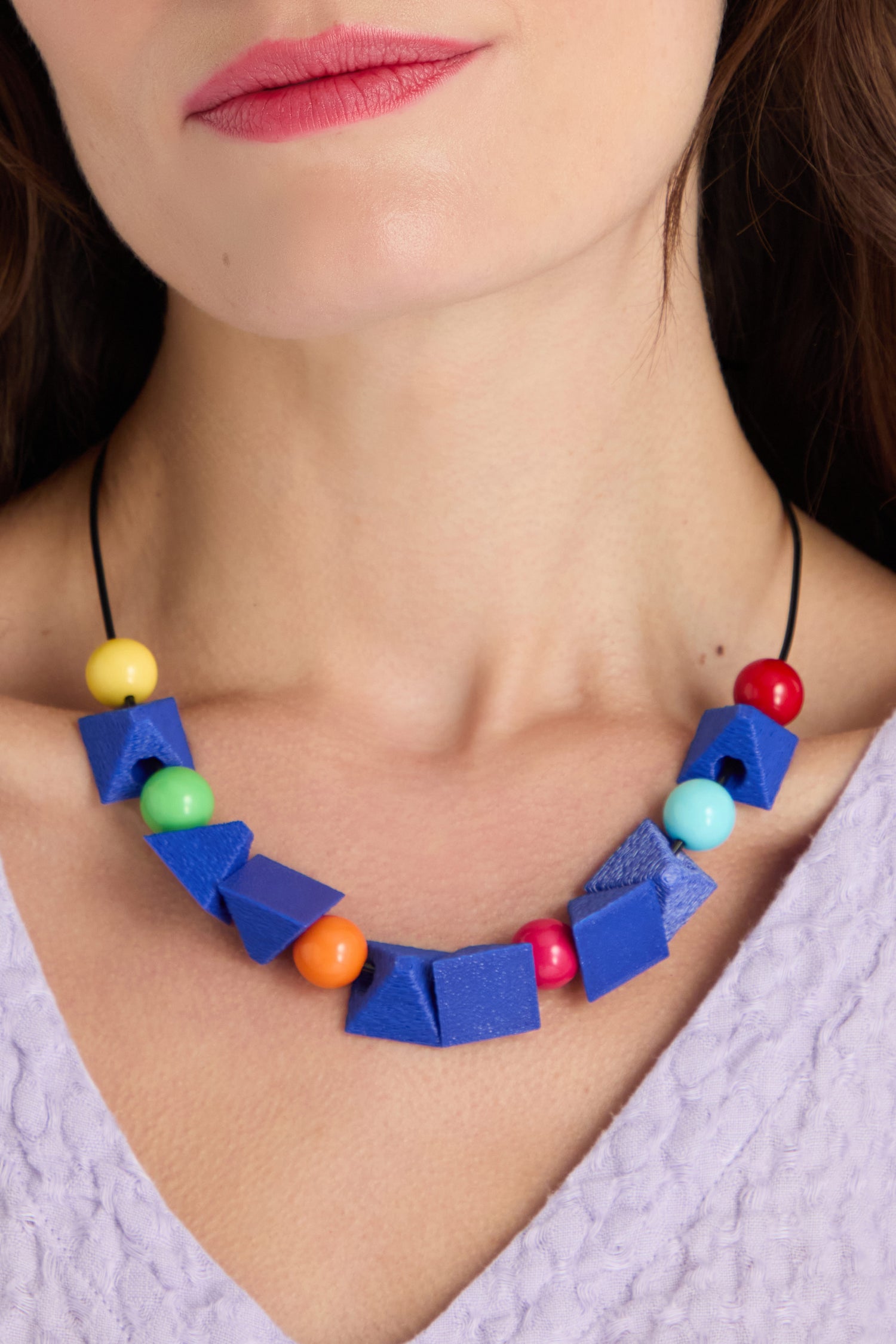 Close-up of a woman wearing the Colour Pop Shapes Necklace, a geometric statement piece with blue and colorful round beads, paired with a light purple textured top. Handmade in Paris, it adds an artistic flair to her outfit.