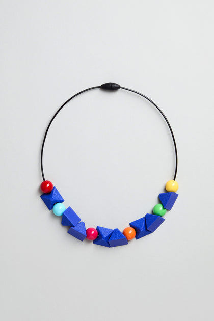 The Colour Pop Shapes Necklace is a handmade geometric statement piece from Paris, with a black wire, large blue beads, and colourful round beads in red, yellow, turquoise, and orange on a plain light background.