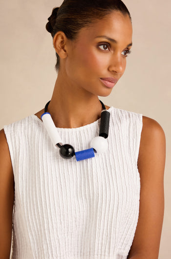 A woman wearing a white textured sleeveless top and the Colour Block Necklace—a contemporary, wearable art piece handmade in France featuring chunky black, white, and blue beads—looks to the side against a neutral background.