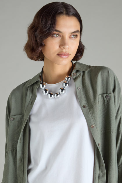 Exuding contemporary elegance, the individual dons a green shirt layered over a white top, accentuated by Samuel Coraux's exquisite 7 Spheres Necklace, as they gaze thoughtfully to the side against a neutral backdrop.