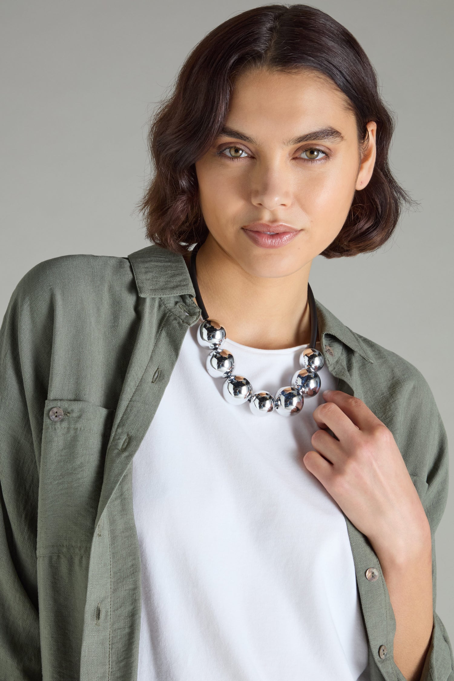 With short brown hair, a person exudes modern sophistication in a green shirt over a white top. The ensemble is elegantly completed with the distinct Samuel Coraux 7 Spheres Necklace, showcasing silver bead artistry.