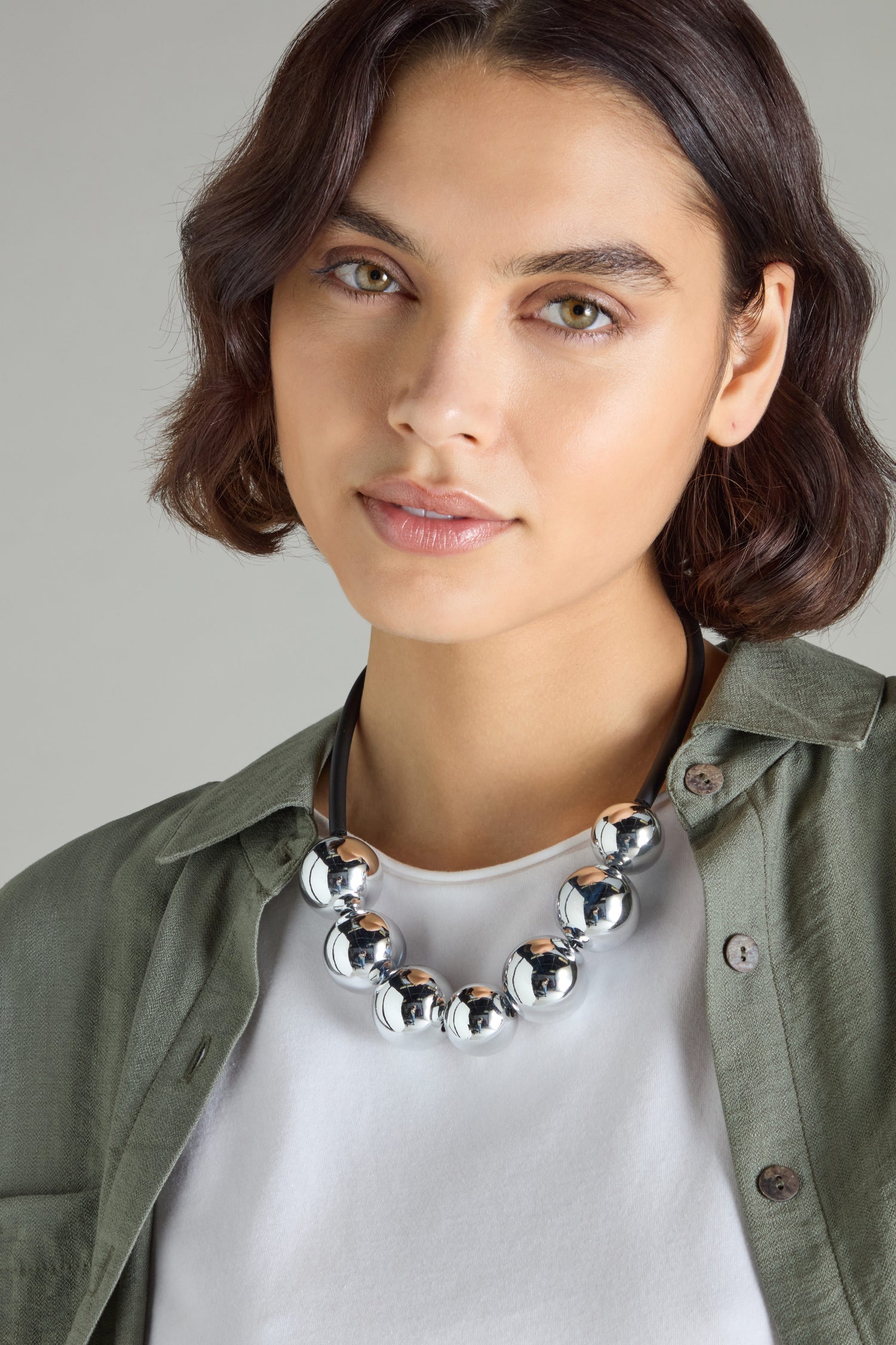 A person with short hair exudes modern sophistication while wearing a green shirt and the striking 7 Spheres Necklace adorned with silver beads, reminiscent of Samuel Coraux's unique designs.