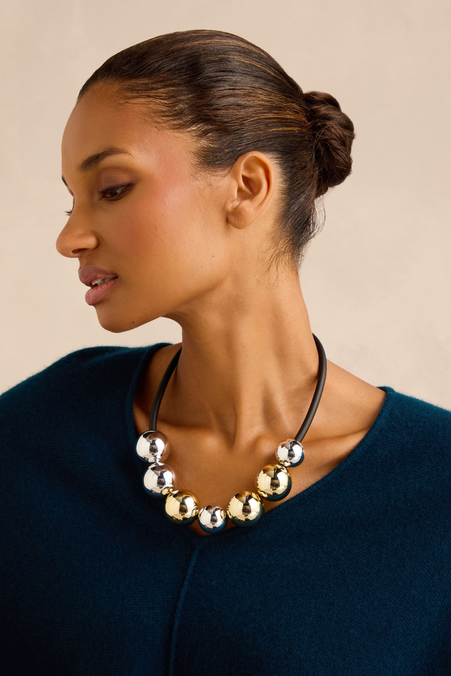 A woman with her hair in a low bun wears a teal top and the Seven Spheres Necklace, a handmade sculptural piece from France featuring alternating large gold and silver beads.