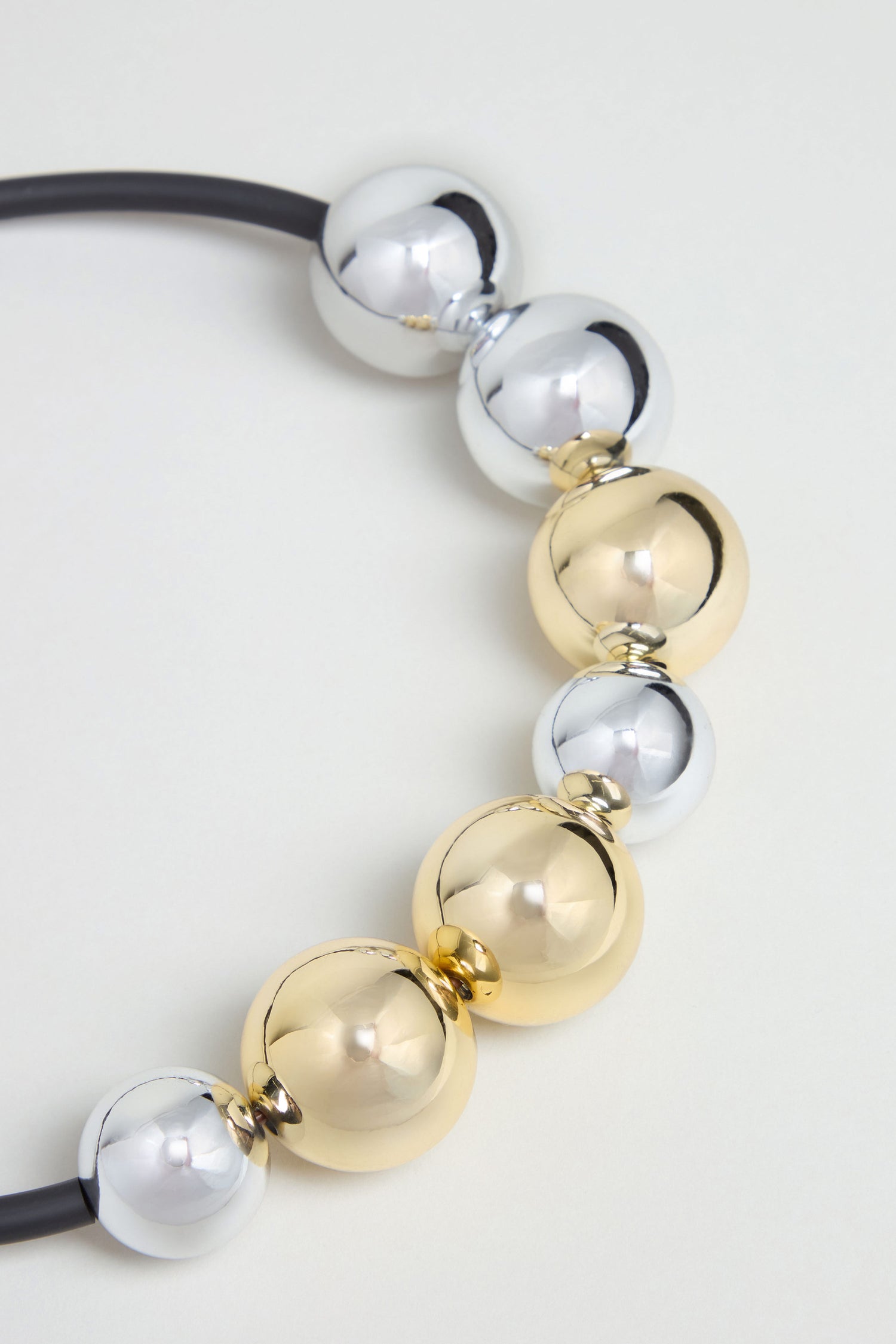 Close-up of the Seven Spheres Necklace, featuring alternating silver and gold metallic spheres strung on a black cord. This sculptural necklace is handmade in France and showcased against a light background.