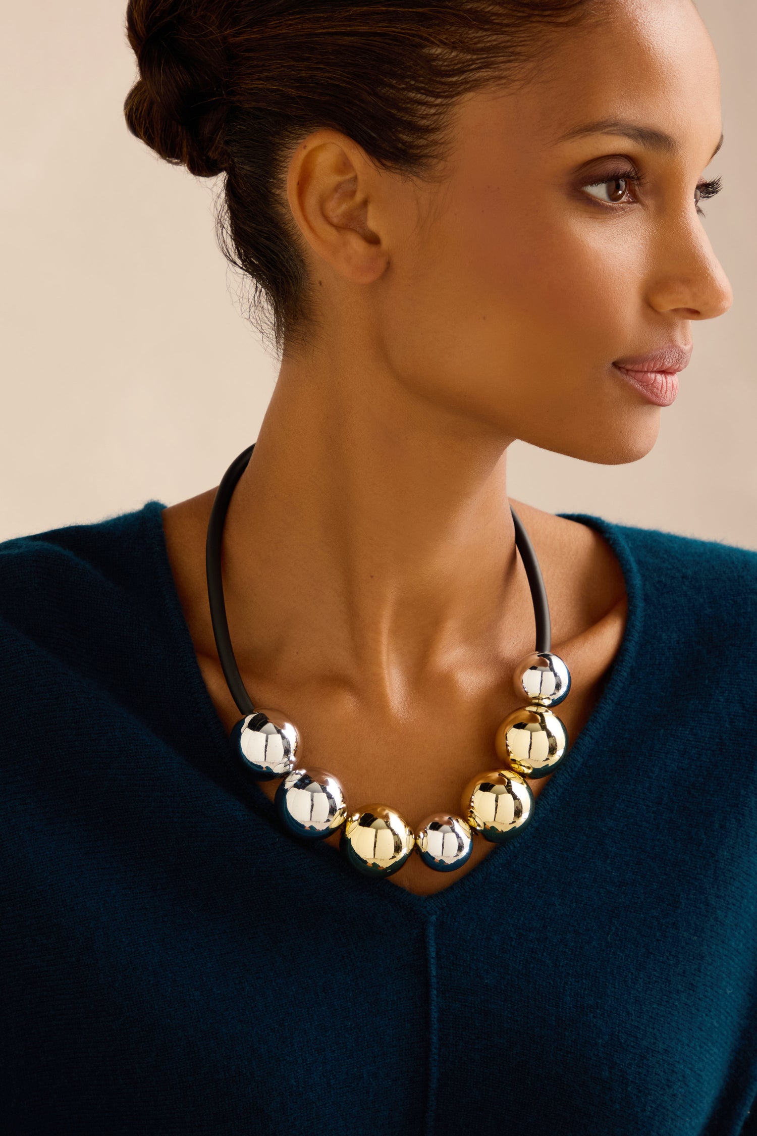 A woman in a teal top gazes to the side against a neutral background, wearing the handmade in France Seven Spheres Necklace, which features large sculptural beads in silver and gold.