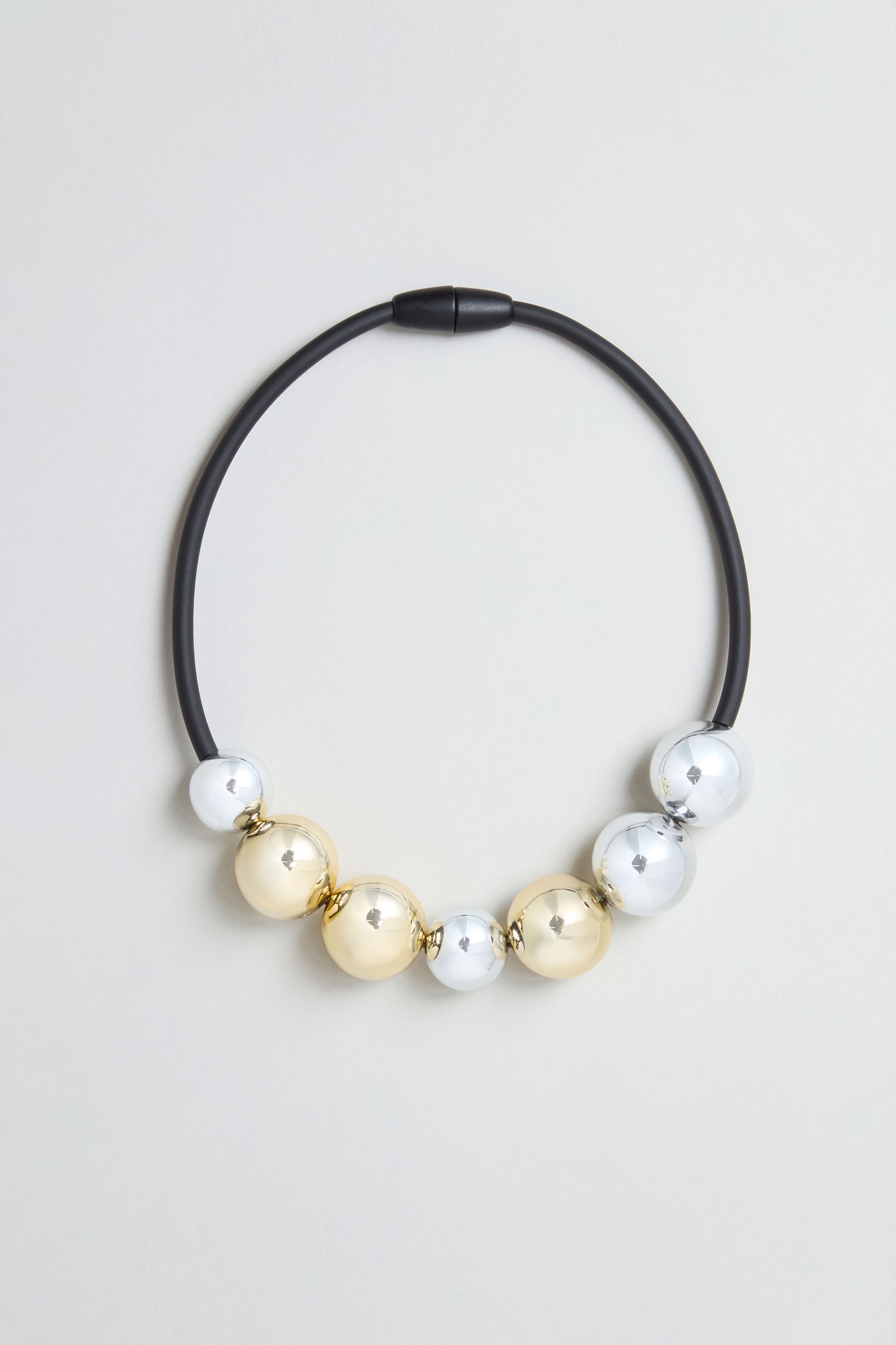 The Seven Spheres Necklace is a sculptural piece, handmade in France, showcasing an eye-catching arrangement of alternating large gold and silver metallic beads against a light gray background.