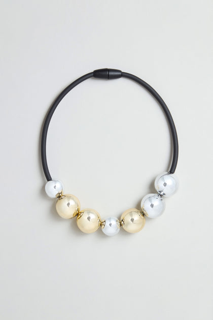 The Seven Spheres Necklace is a sculptural piece, handmade in France, showcasing an eye-catching arrangement of alternating large gold and silver metallic beads against a light gray background.