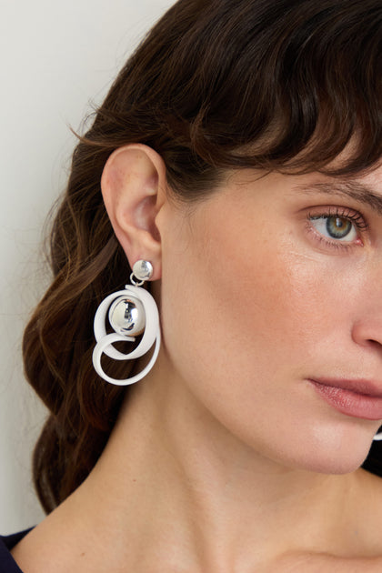 Close-up of a woman with shoulder-length brown hair wearing Silver Sphere Penseur Earrings, which showcase white and silver circular shapes. The lightweight design enhances the modern appeal of these earrings.