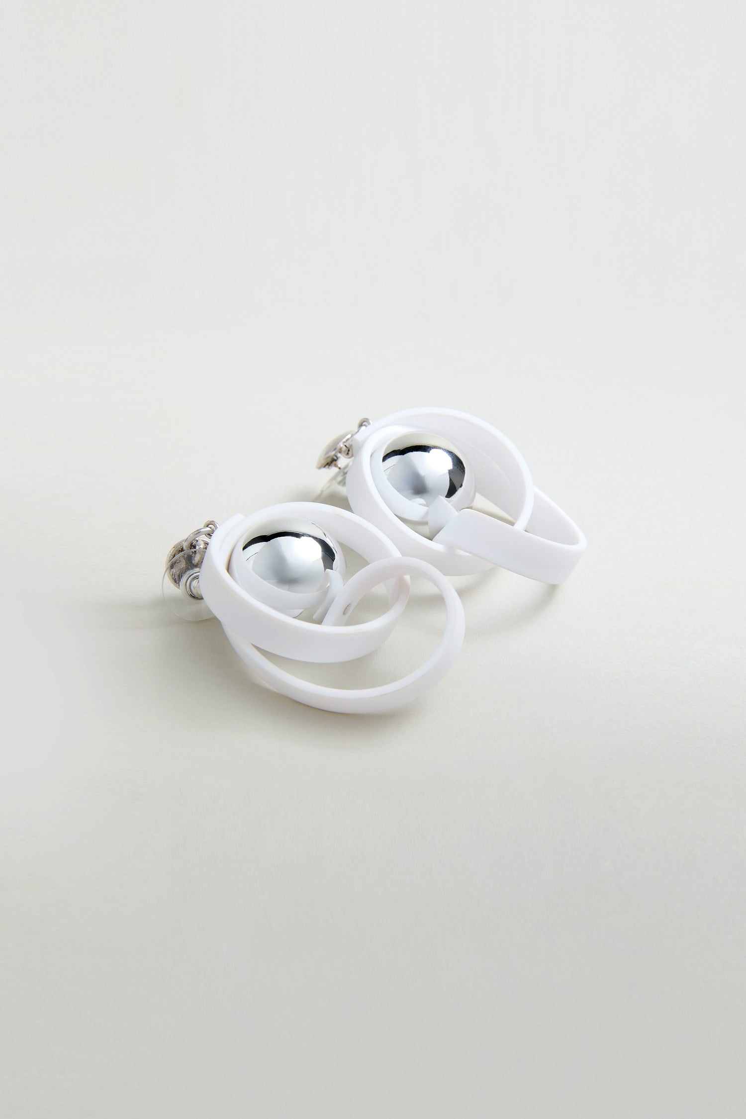 The Silver Sphere Penseur Earrings feature shiny silver spheres wrapped in overlapping white loops with a modern, lightweight design, shown against a light, neutral background.