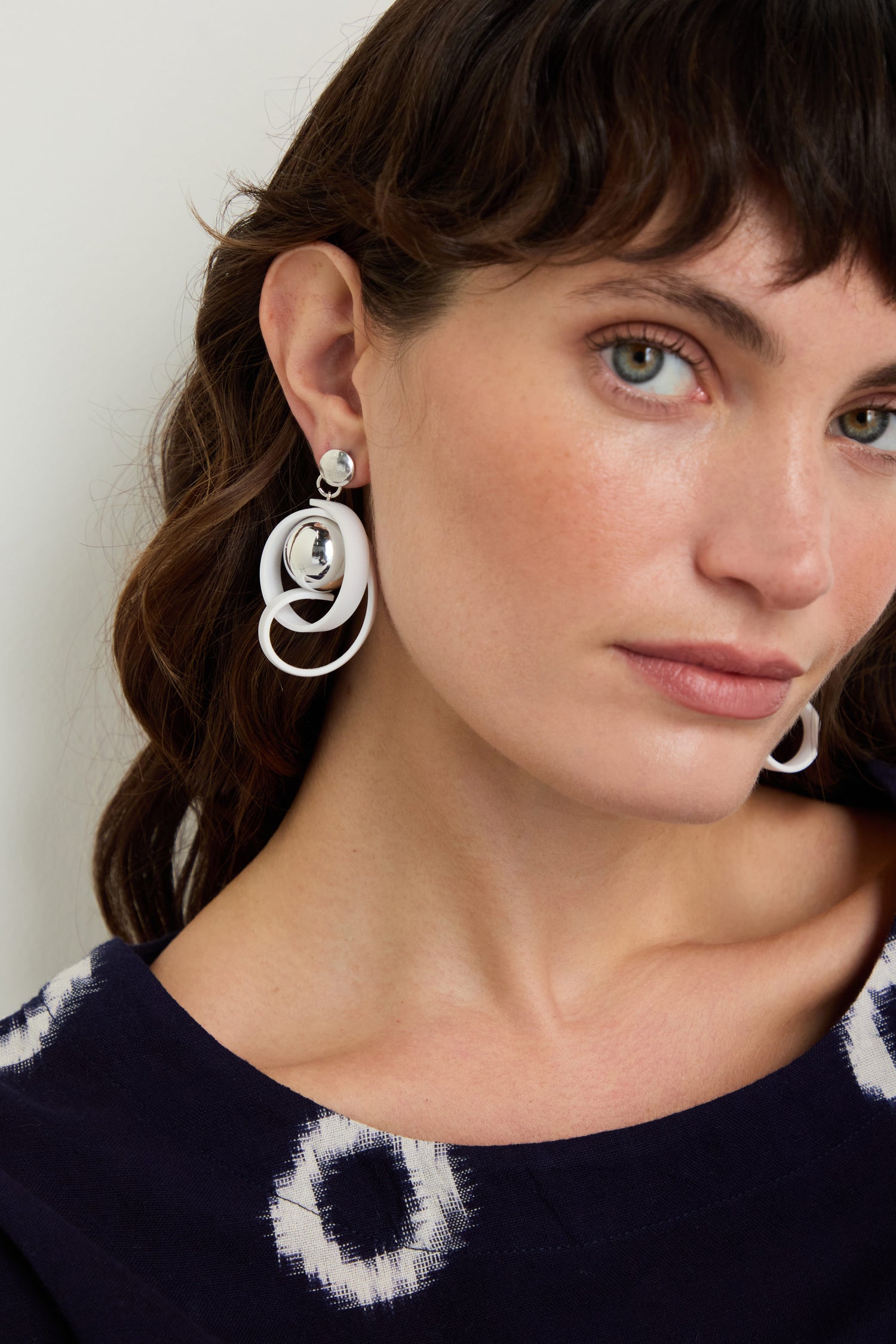 Woman with wavy brown hair wears a navy blue top with white circular patterns and Silver Sphere Penseur Earrings, posing against a plain background.
