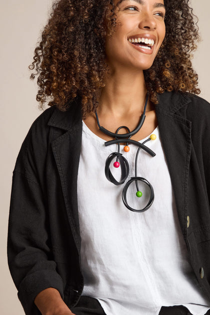 A woman with curly hair is smiling, wearing a black jacket over a white shirt, and a Space Necklace adorned with large, colorful beads, reminiscent of Samuel Coraux jewelry.